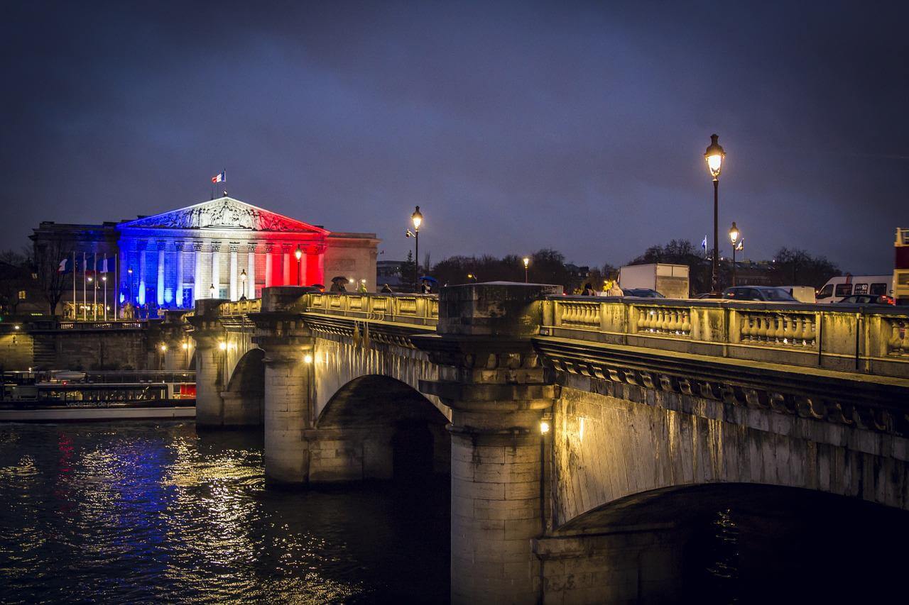 French flag colors projected onto a French building overlooking a river and bridge at night