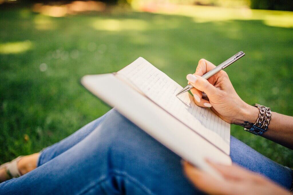 A woman writing on a notebook in an open field of green grass.