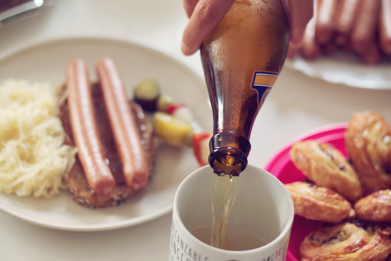 A German beer being poured in a ceramic mug. Two sausages sit on a plate in the bakcground.