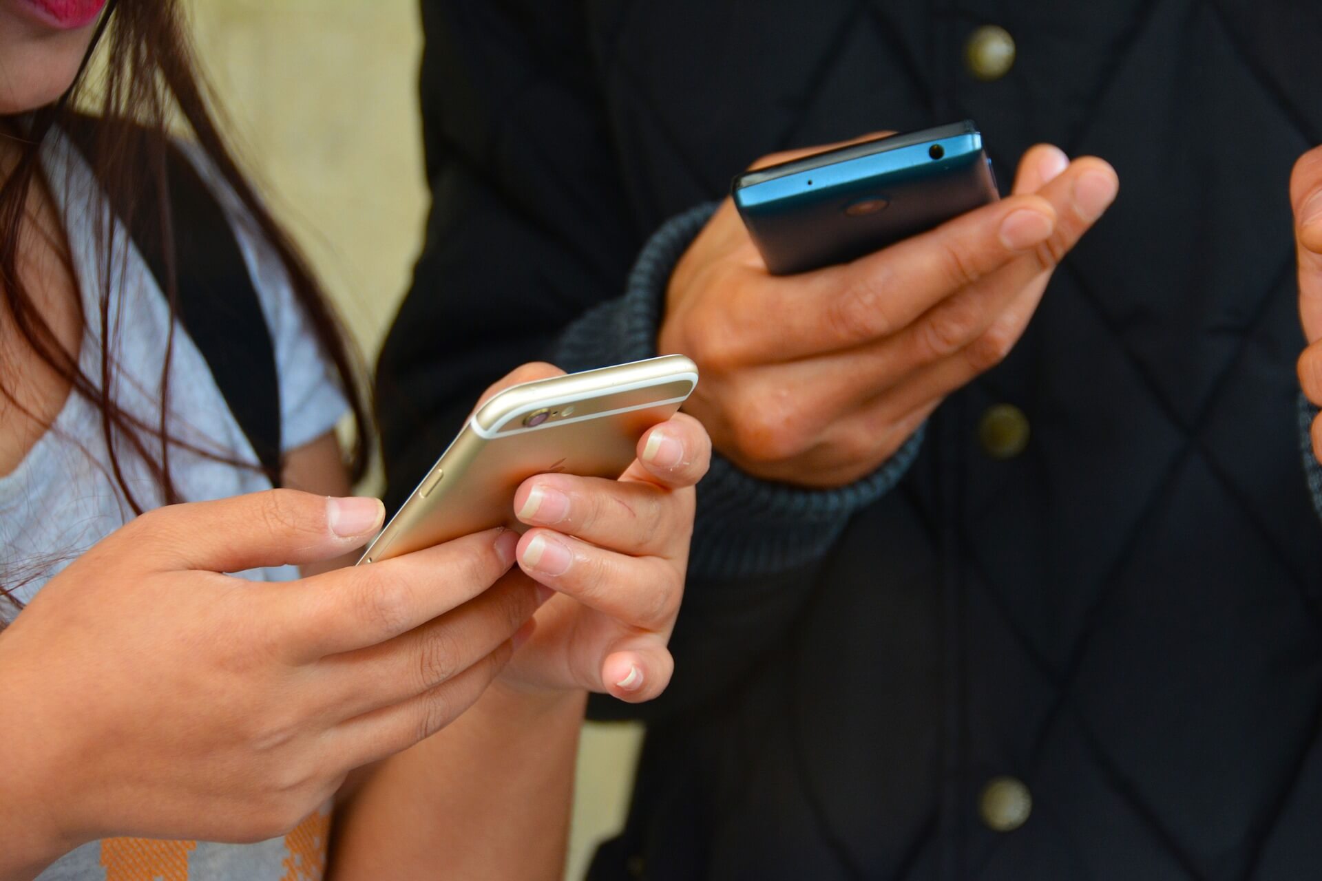Two people stand next to each other with their smartphones in their hands. One of them, a young woman, holds hers with both hands, while the other, a man, holds his with one.