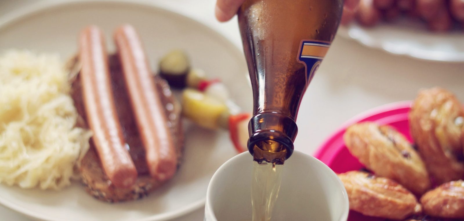 A German beer being poured in a ceramic mug. Two sausages sit on a plate in the bakcground.